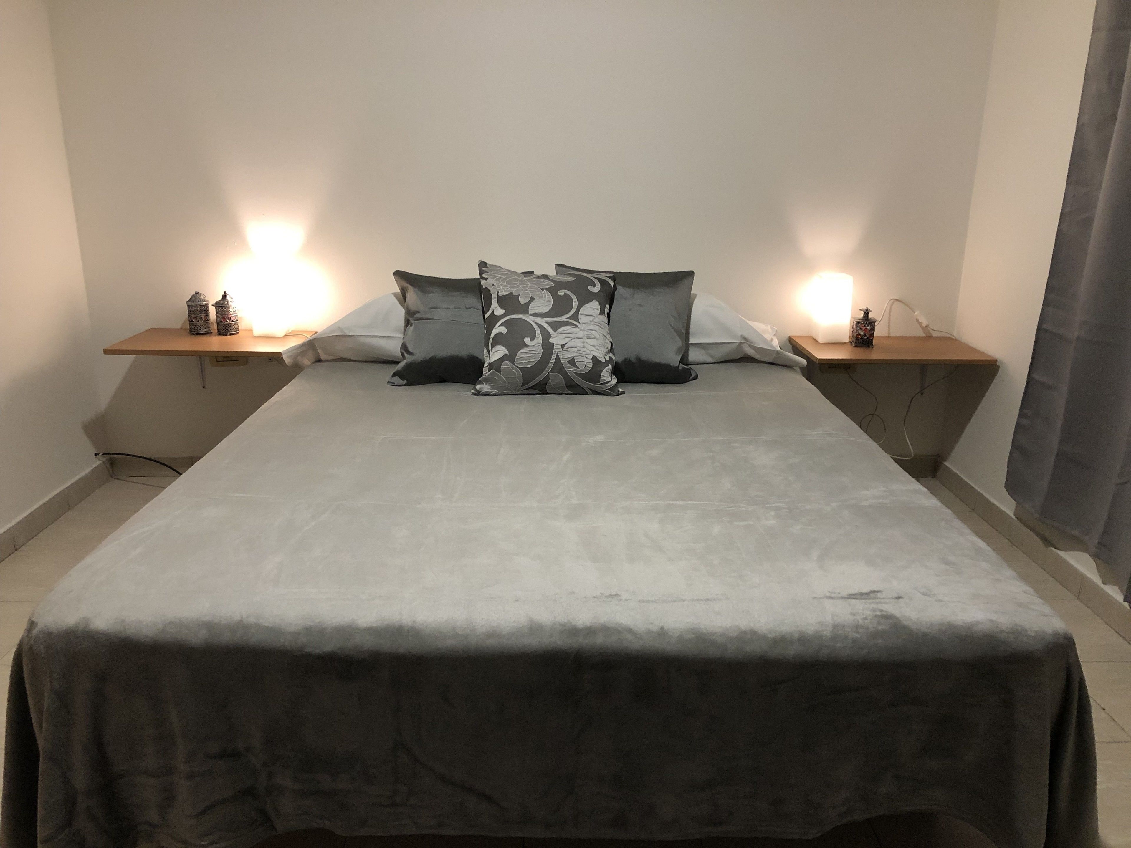 Una cama bien hecha con una colcha gris y dos almohadones decorativos, con mesitas de noche a cada lado que tienen lámparas y pequeños adornos.