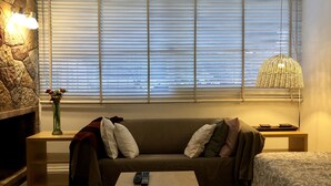 Ein gemütliches Wohnzimmer mit Kamin aus Stein, einer Couch mit Kissen, einem Couchtisch mit Fernbedienungen und einem Fenster mit Jalousien.