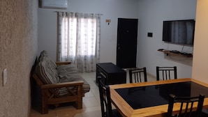 A living room with a sofa, a wooden dining table with chairs, a TV mounted on the wall, and a black cabinet.