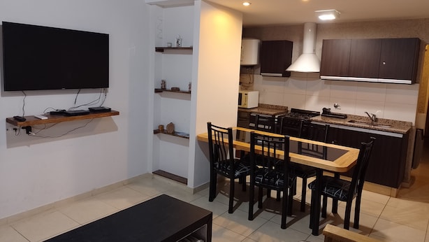 A compact living space with a TV mounted on the wall, a dining area with a wooden table and chairs, and a kitchen area with dark cabinets and a stove.