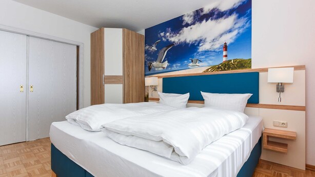 A hotel room with a large bed, a painting of a lighthouse and seagulls, a wooden wardrobe, and a nightstand with a lamp. - Vacation apartment Seestern - Holiday apartments Der Seehund, Haus Kajedeich in (Esens)