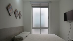 A bedroom with a bed, a window with blinds, and a flat-screen TV mounted on the wall. - Apartamentos MED (Medellín)
