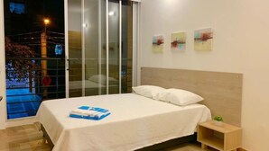A bedroom with a bed, a nightstand, a small table, and a window with a view of the outside. - Apartamentos MED (Medellín)