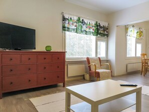 Una sala de estar con televisor, un aparador rojo, una silla, una mesa blanca y una ventana con persianas.