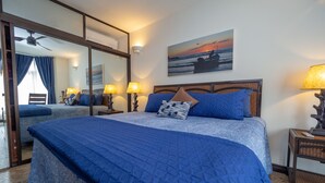 A bedroom with a large bed, a ceiling fan, a nightstand with a lamp, and a painting of a sunset over the ocean. - Luxury Beachfront Mar Arena 2 (Jacó)