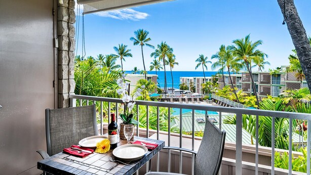 A balcony with a table set for two, overlooking a pool and palm trees. - 🏖️Beautiful Ocean Views | Top Floor | Concierge Included (Kailua Kona)