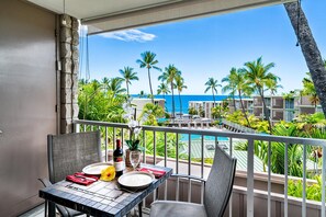 A balcony with a table set for two, overlooking a pool and palm trees. - 🏖️Beautiful Ocean Views | Top Floor | Concierge Included (Kailua Kona)