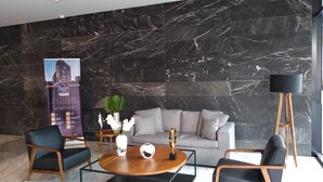 A modern lobby with a black marble wall, a grey sofa, two armchairs, a coffee table, a floor lamp, and a poster advertising a hotel.