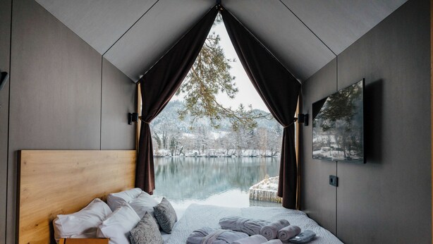 Une chambre moderne avec une tête de lit en bois, un lit recouvert d’une couette et de draps blancs et gris, une pile de serviettes enroulées, une petite table de chevet en bois et une vue sur un lac, visible à travers une fenêtre triangulaire.