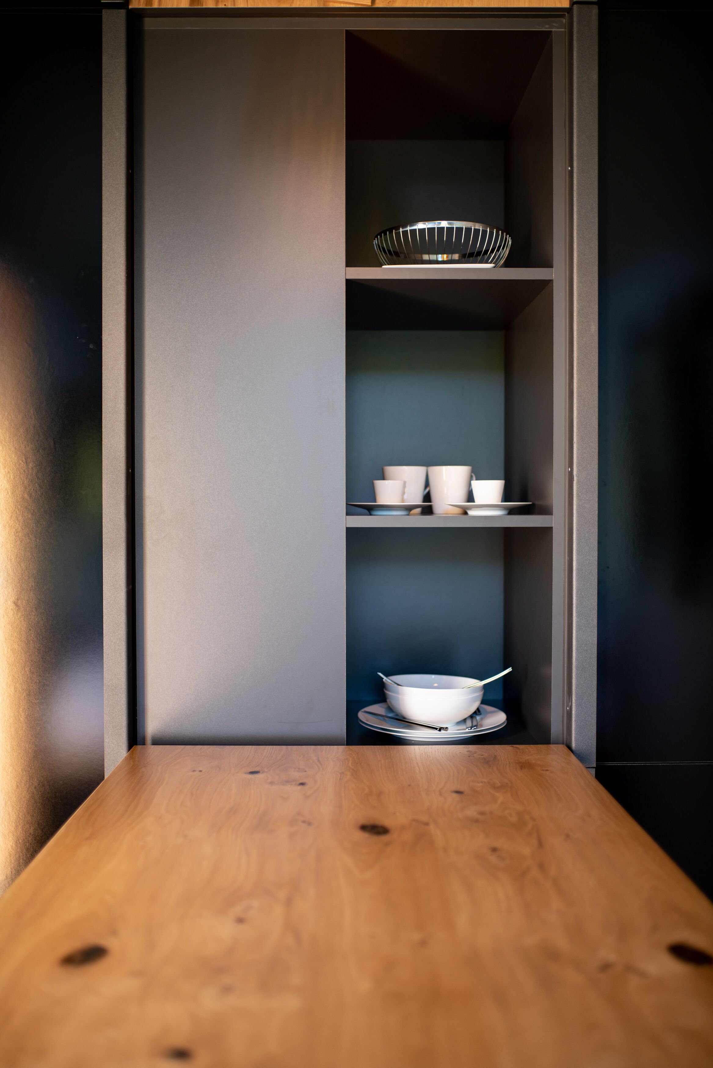 A kitchen with a wooden countertop, a metal bowl on the shelf, and white cups and bowls on the lower shelf.