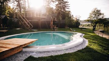 A swimming pool with wooden deck seating, surrounded by a grassy area and trees.