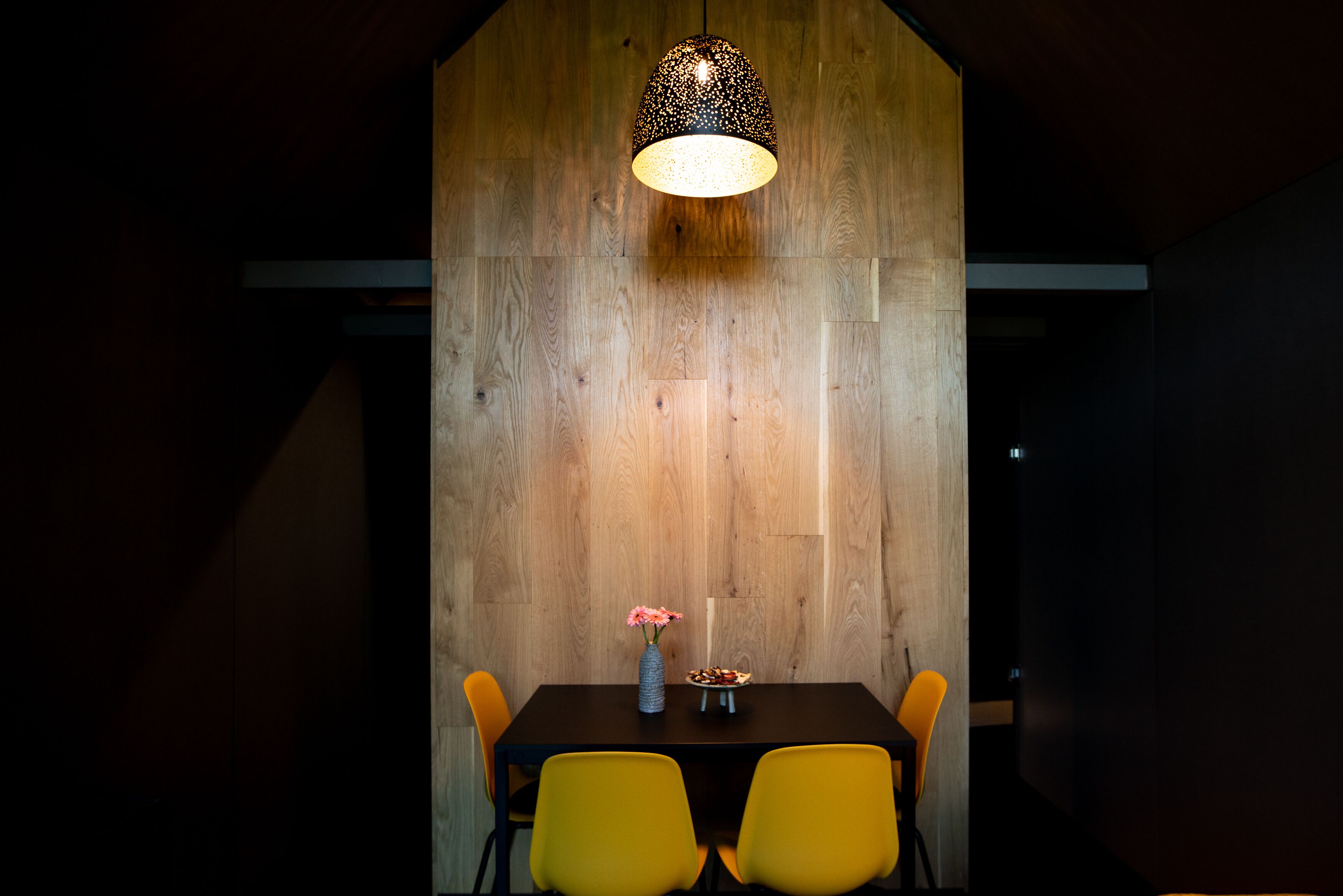 Ein Essbereich mit Holzwand, einer Hängelampe, einer Blumenvase und gelben Stühlen.