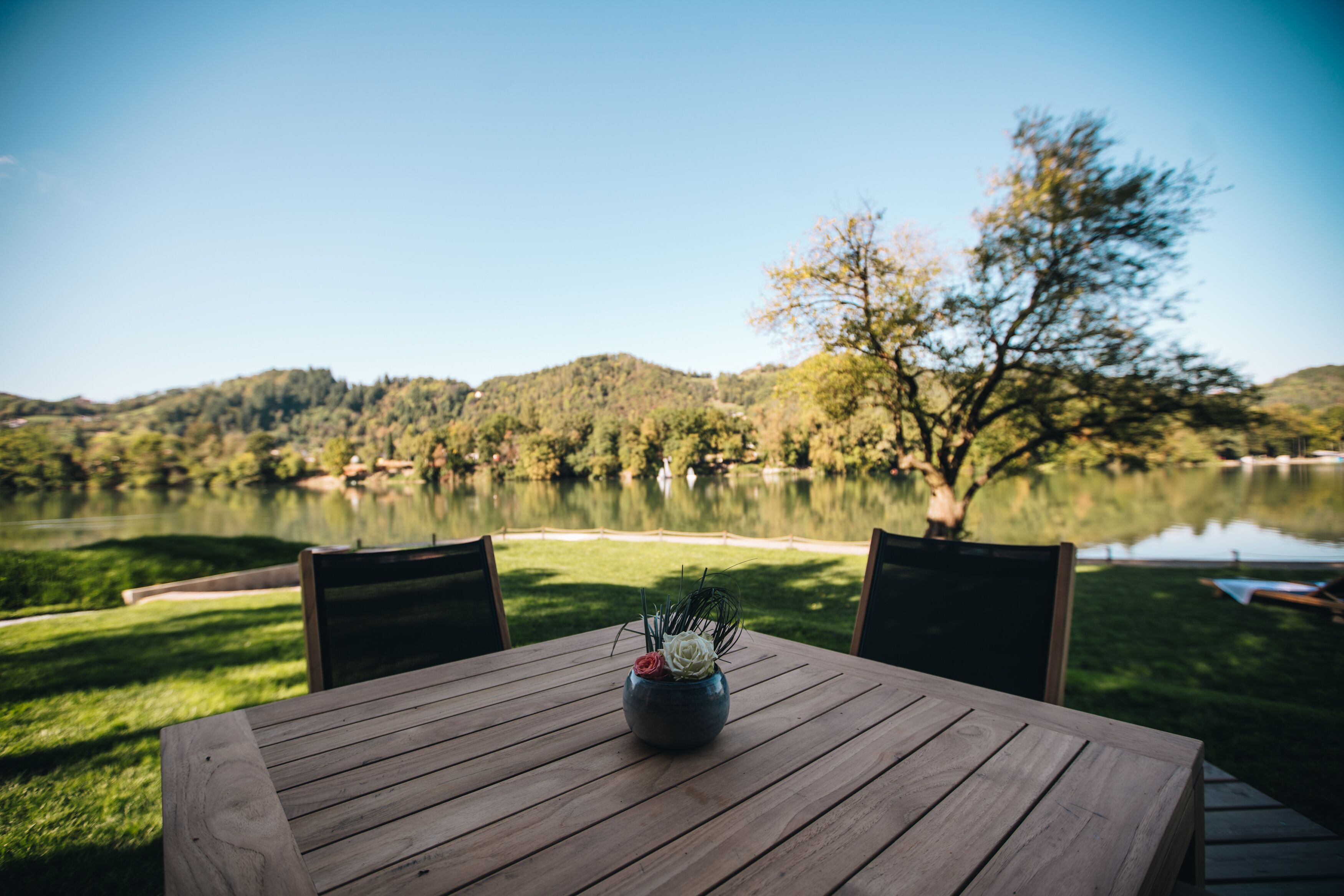 Una mesa de madera con un florero, dos sillas y vista a un lago y colinas.