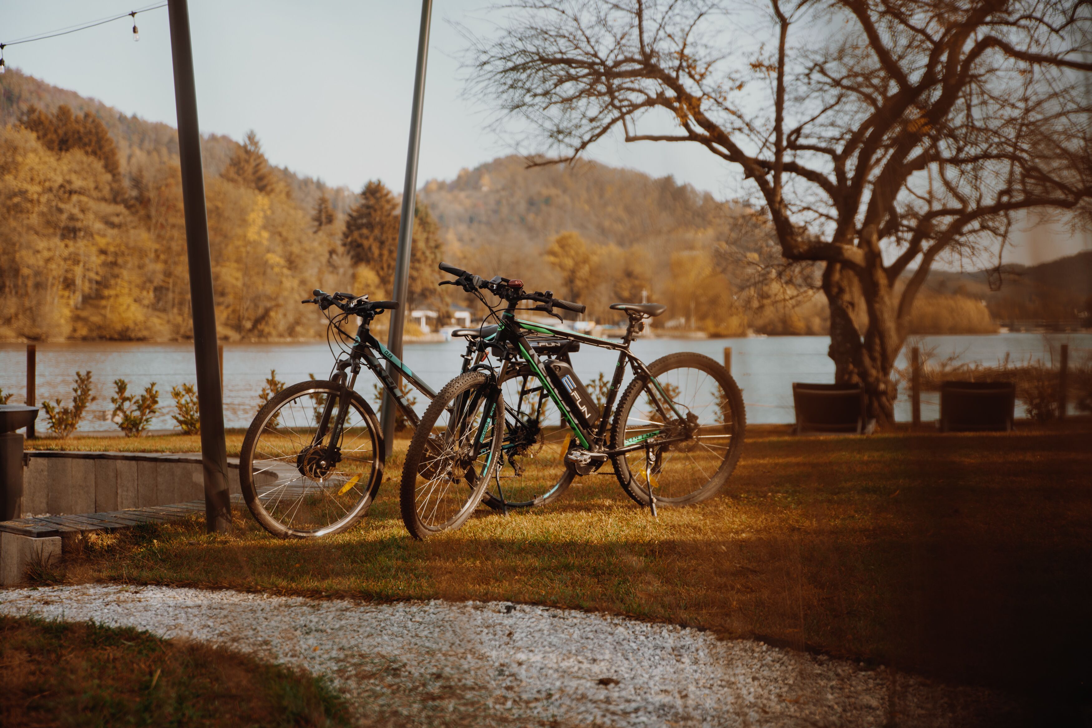 Two bicycles parked on a gravel path near a lake with trees and hills in the background.