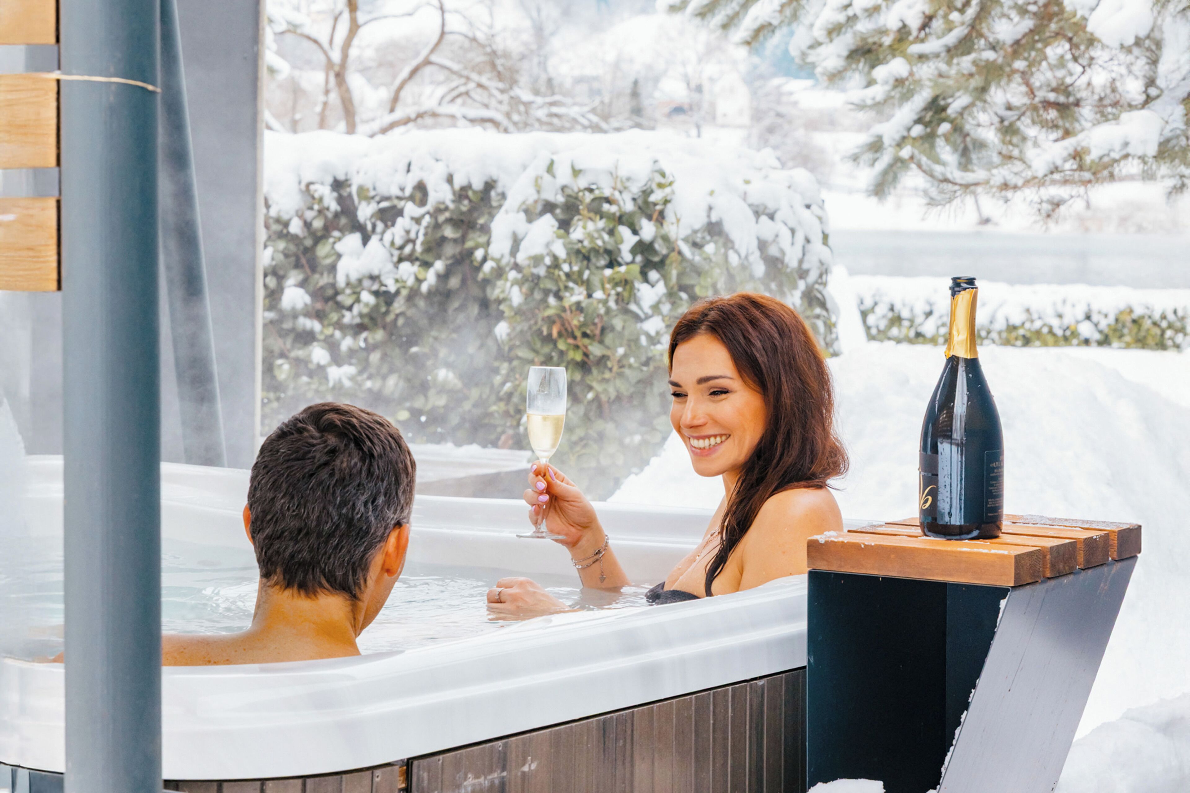 A hot tub with a bottle of champagne and a glass, surrounded by snow-covered trees.
