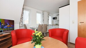 A room with a wooden dining table, two red chairs, a yellow flower vase, a television, and a kitchen area in the background.