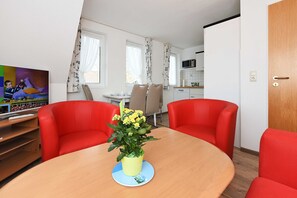 Ein Zimmer mit Esstisch aus Holz, zwei roten Stühlen, einer gelben Blumenvase, einem Fernseher und einer Küchenzeile im Hintergrund.