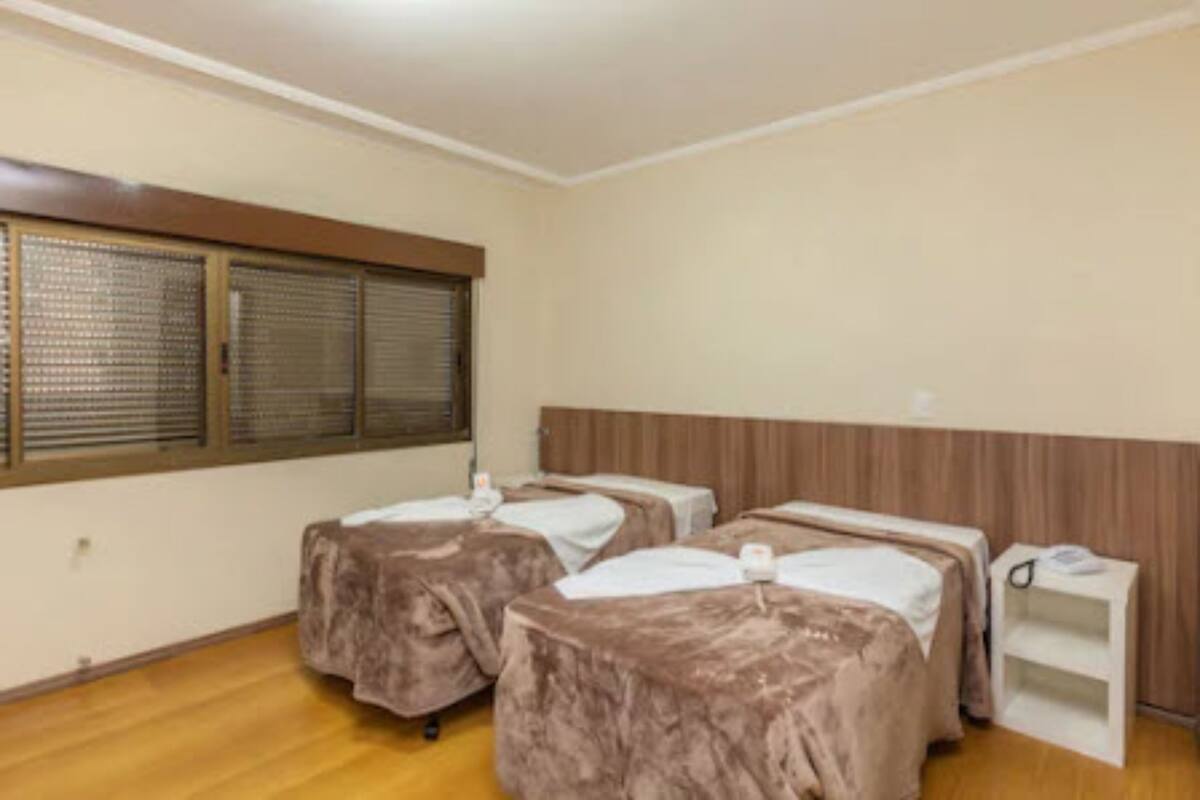 A room with two beds, a wooden headboard, a window with blinds, and a white nightstand.