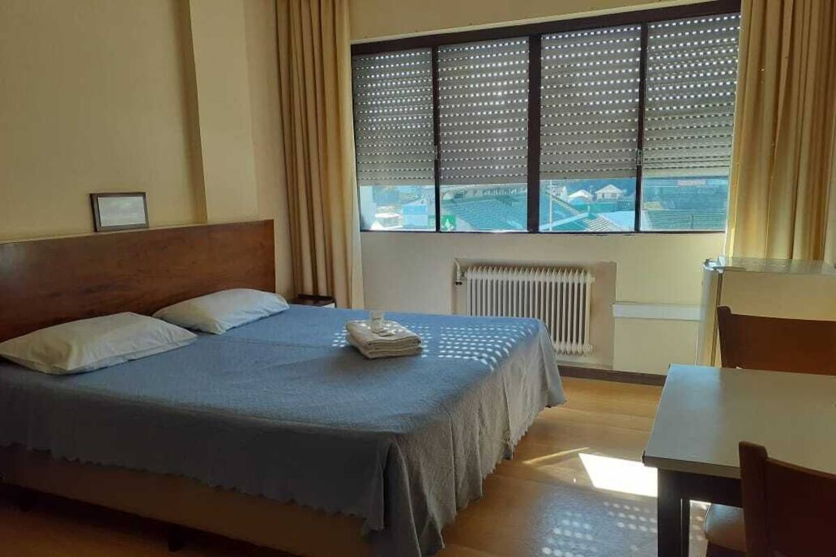 A hotel room with a bed, a table, a window with blinds, and a radiator.