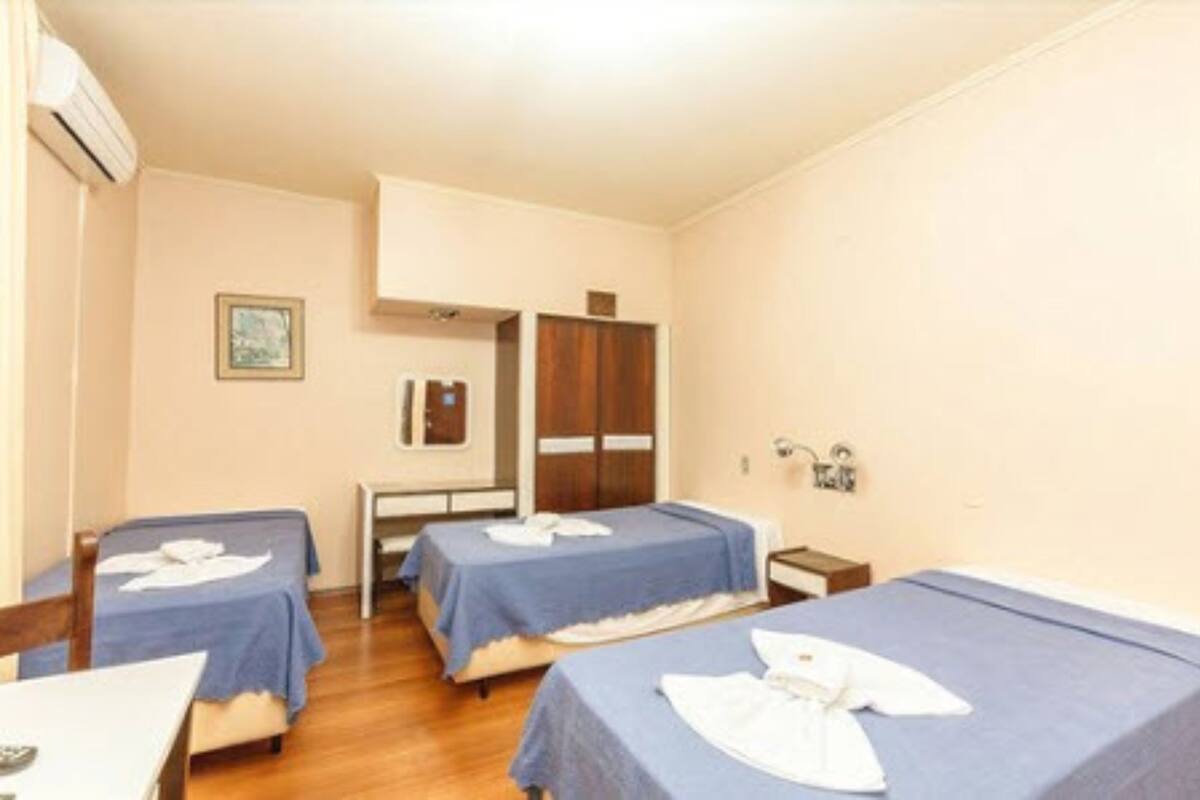 A hotel room with two single beds, a small table, a chair, a wardrobe, and a framed picture on the wall.