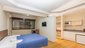A bedroom with a bed, a window with blinds, a small desk, and a kitchenette area with a microwave and a sink.