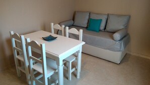 Une petite pièce, bien rangée, avec une table et des chaises blanches, un canapé gris agrémenté d’un coussin turquoise et un sol carrelé.