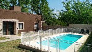 A rectangular swimming pool with a white safety fence, surrounded by a grassy yard and a building with a brown exterior.