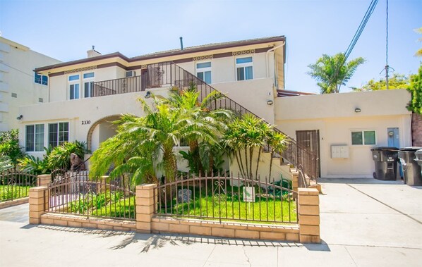 A two-story building with a balcony, a gated entrance with palm trees, and a clear sky.