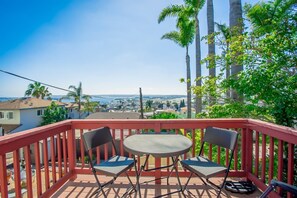 A balcony with two chairs and a table, overlooking a coastal town with palm trees and houses.
