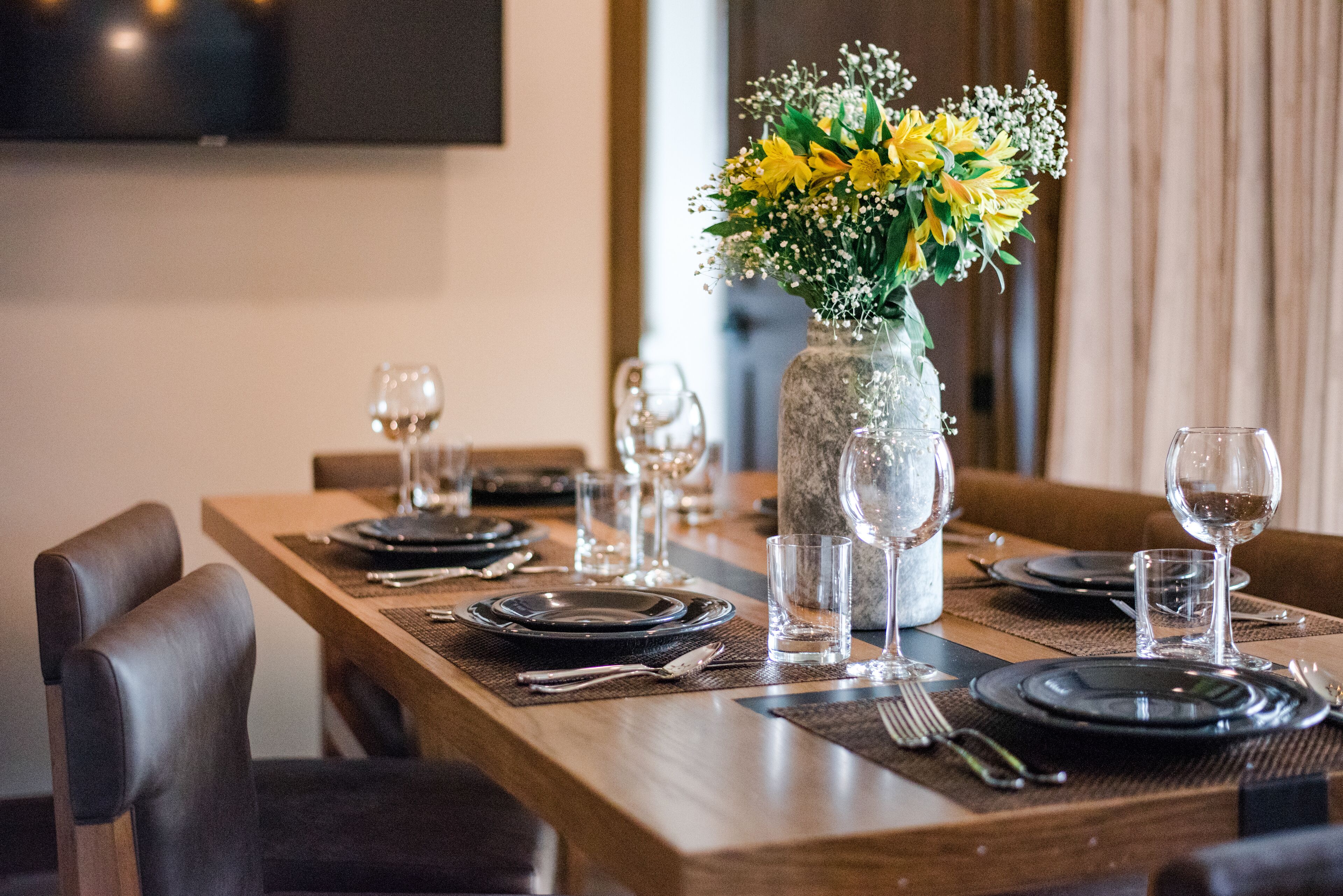 A dining table set with plates, glasses, and cutlery, with a vase of yellow flowers in the center.