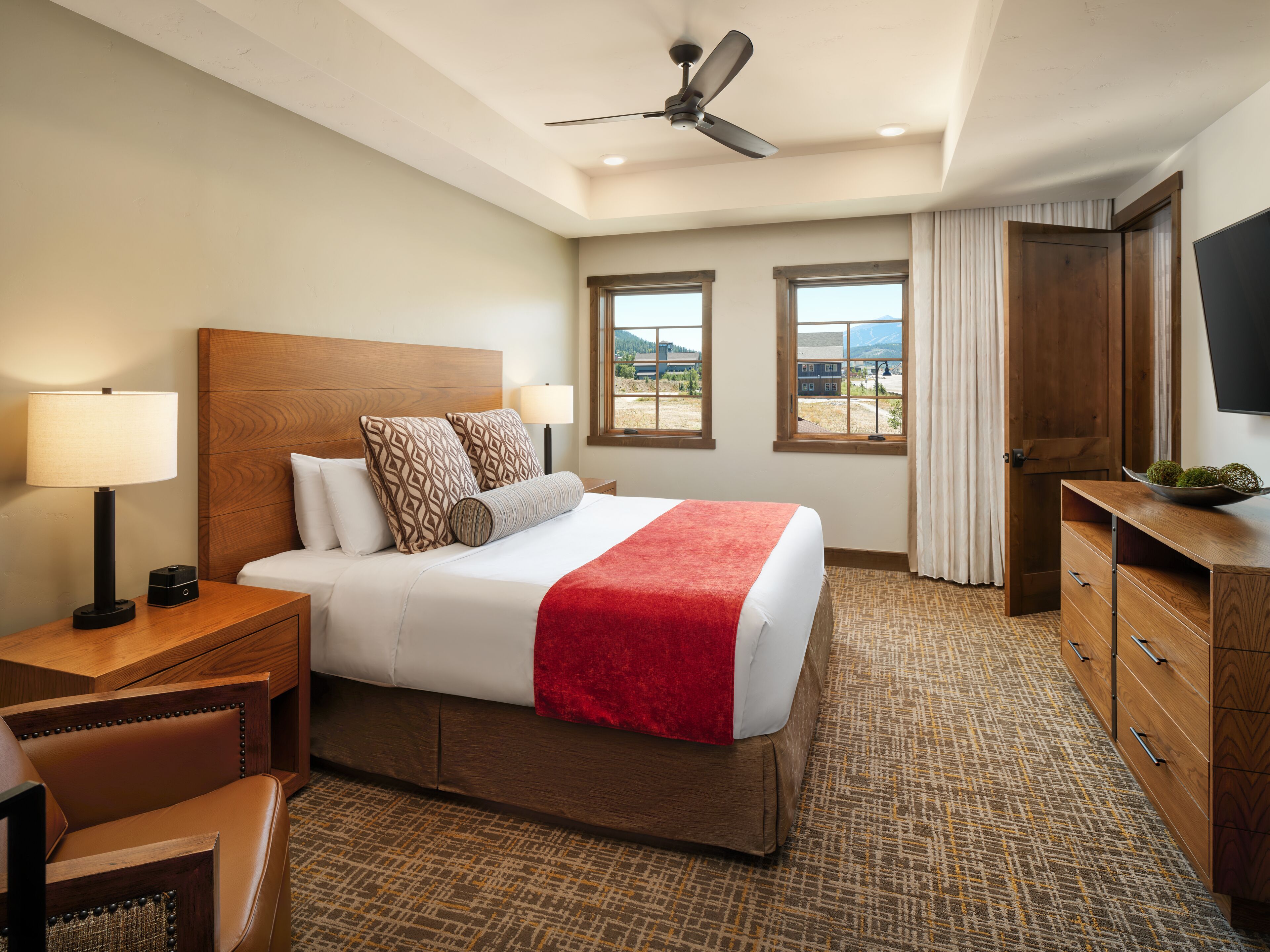 A hotel room with a large bed, a wooden headboard, two bedside lamps, a desk with a lamp, a television, and a view of mountains through the windows.
