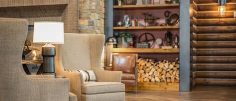 A cozy living room with a fireplace, a comfortable armchair, a wooden bookshelf, and a log storage area.