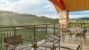 Una terraza de madera con mobiliario exterior, un pilar de piedra y vistas a un lago y colinas.