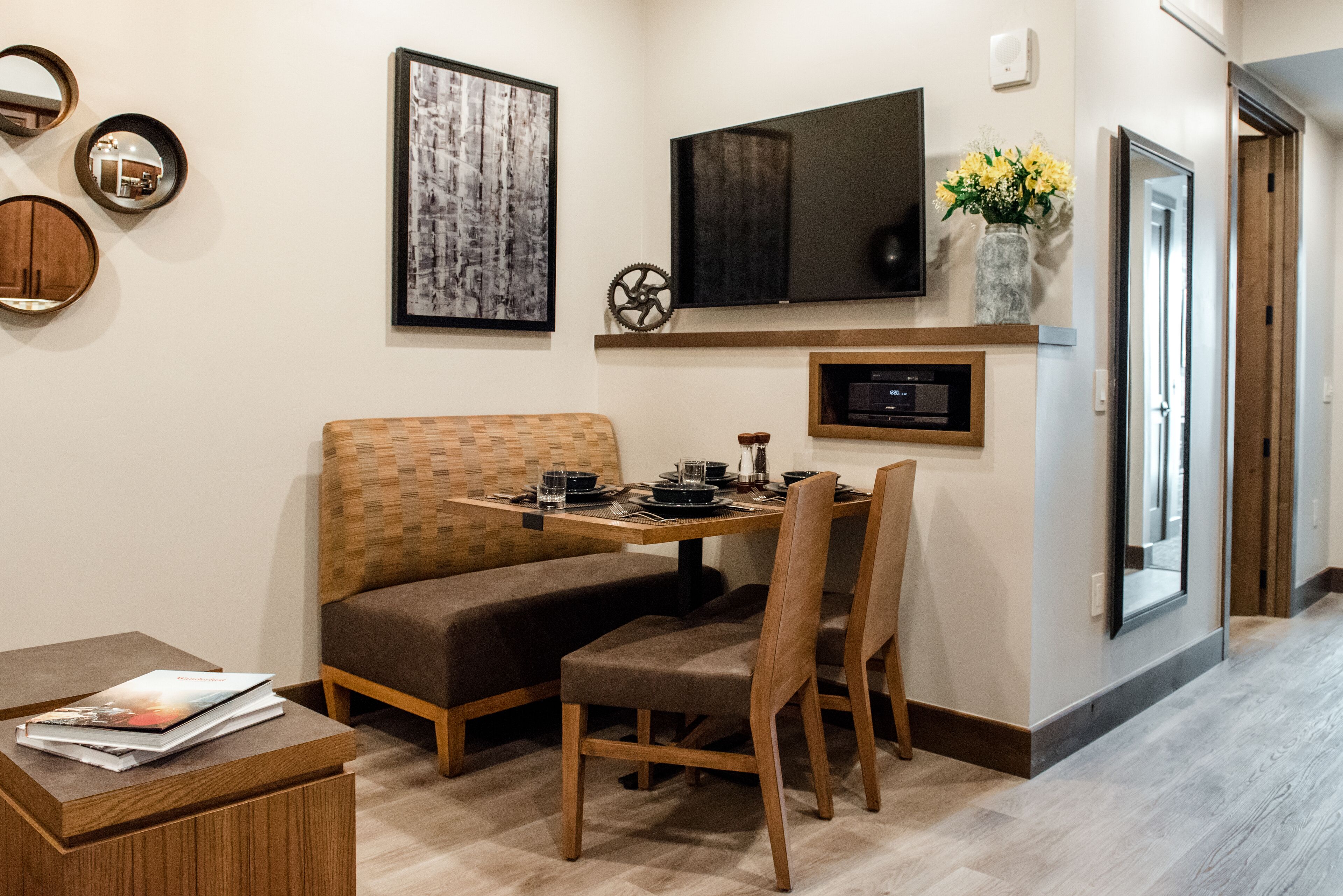 Un coin repas avec une table et un banc en bois, un téléviseur à écran plat fixé au mur juste au-dessus, et un vase avec des fleurs posé sur le mur.