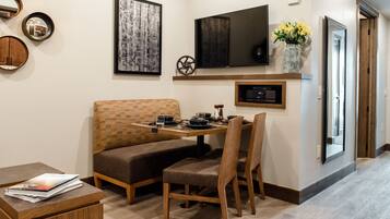 Un coin repas avec une table et un banc en bois, un téléviseur à écran plat fixé au mur juste au-dessus, et un vase avec des fleurs posé sur le mur.