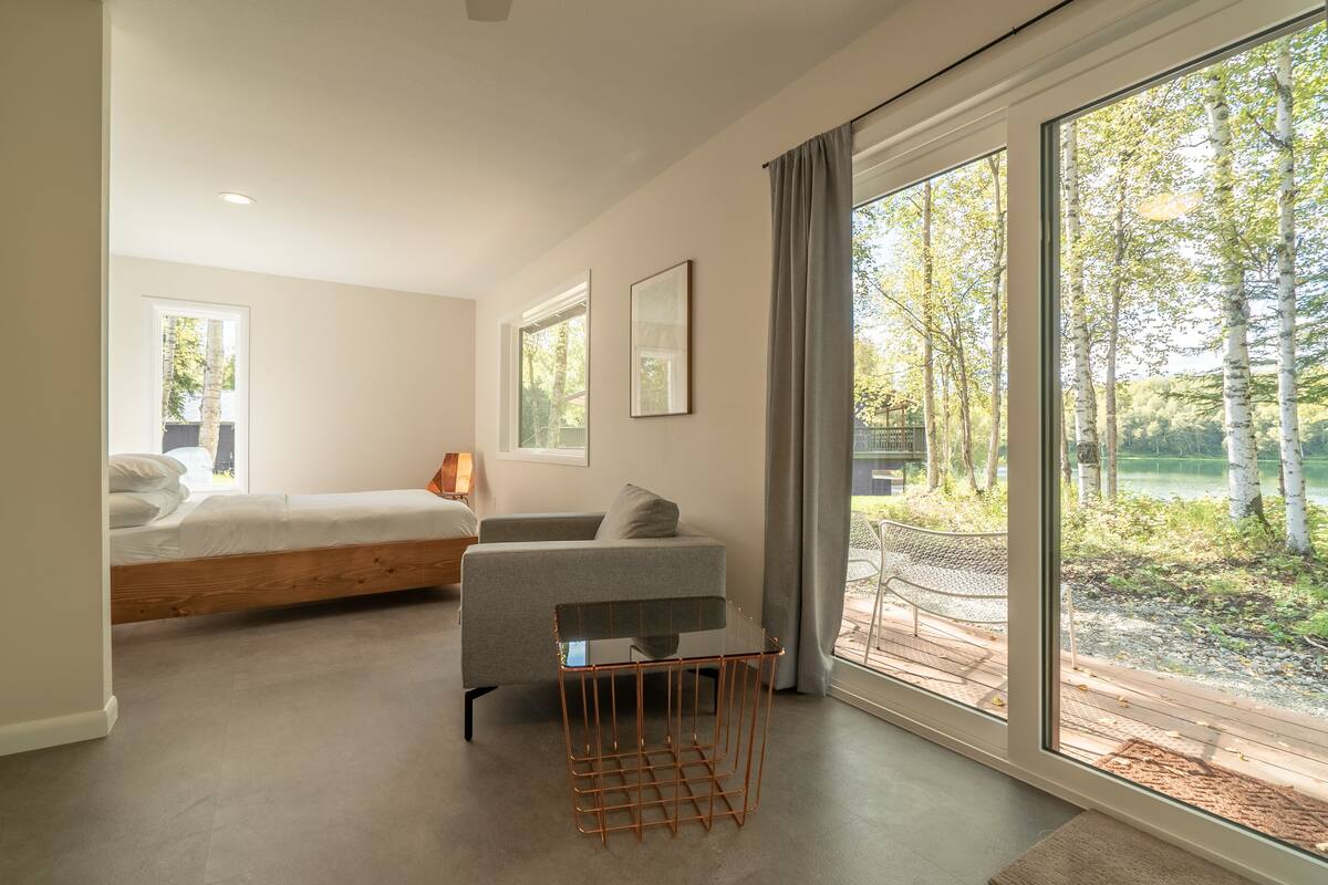 Un dormitorio moderno con cama, sofá, una mesita de centro de vidrio y ventanas grandes que dan a vistas de árboles y un lago.