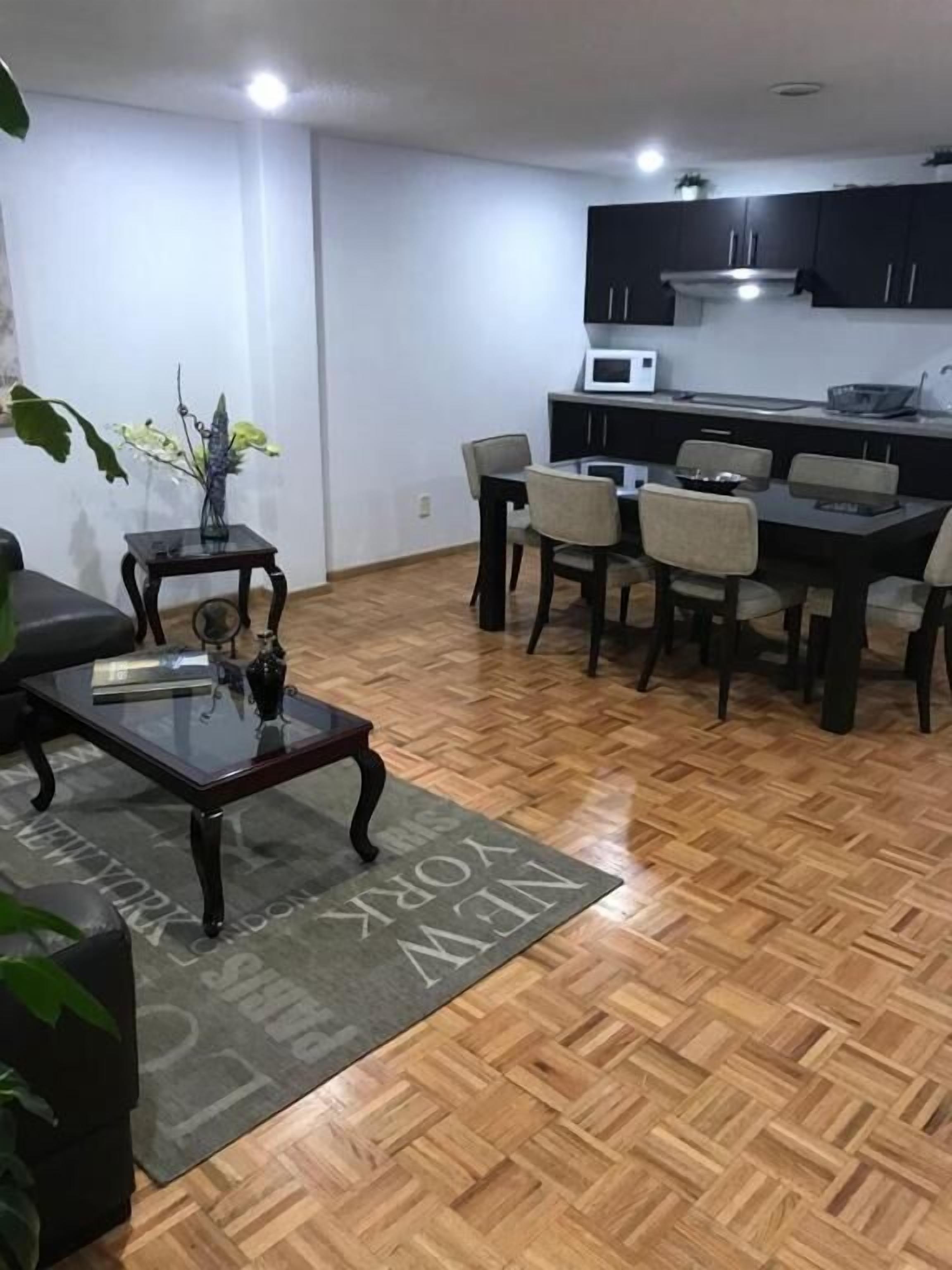 Una sala de estar moderna con comedor, piso de parqué de madera, y cocina con armarios oscuros y un microondas.