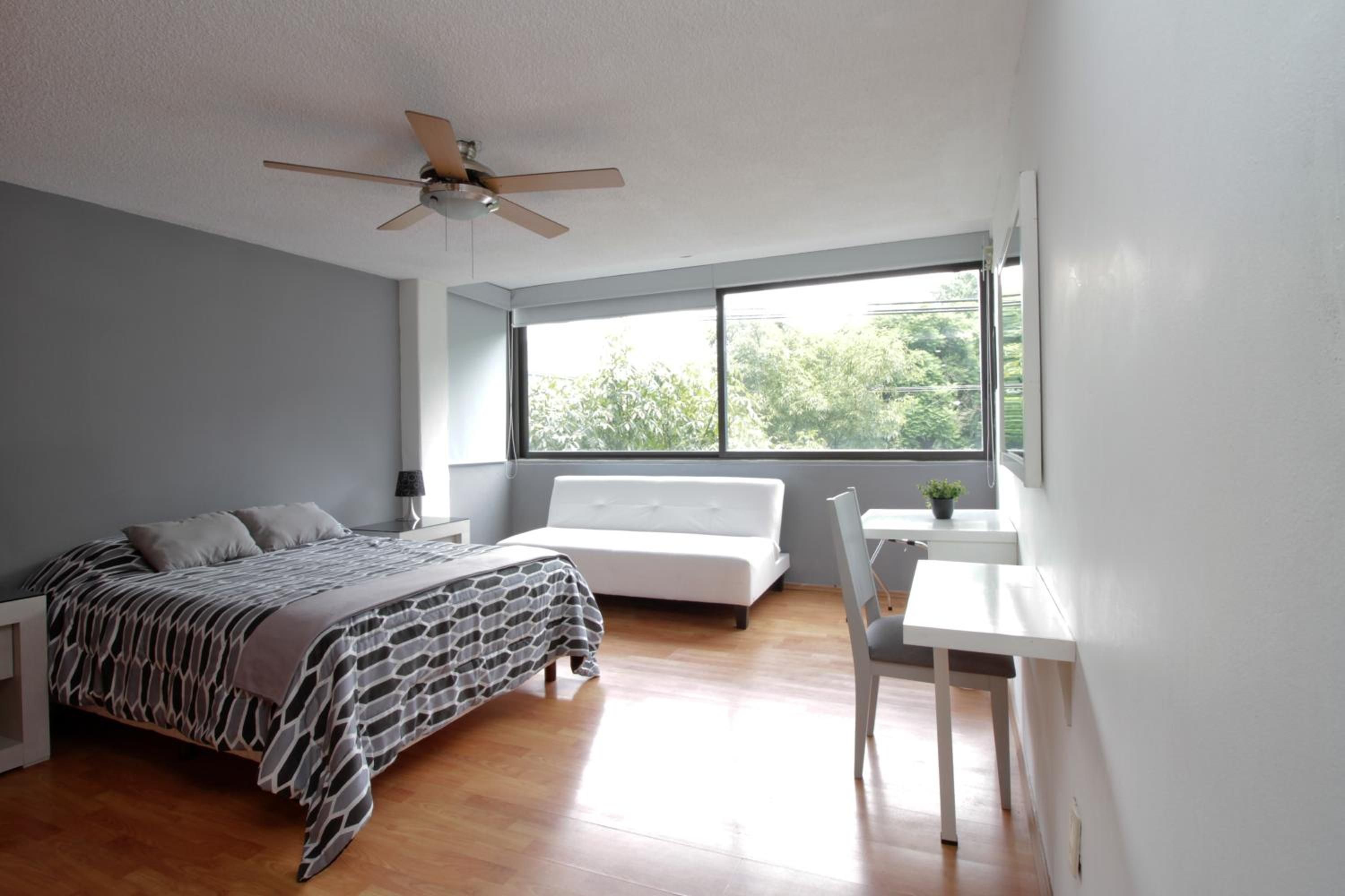 Un dormitorio con una cama, un escritorio, una silla, un ventilador de techo y un ventanal grande.