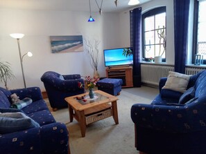 A living room with blue patterned sofas, a wooden coffee table, a flat-screen TV, a painting of a beach, and a large window with curtains.