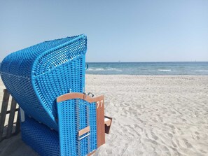 A beach chair with a metal frame and a wooden seat, positioned on a sandy beach with a clear view of the ocean and a distant sailboat.