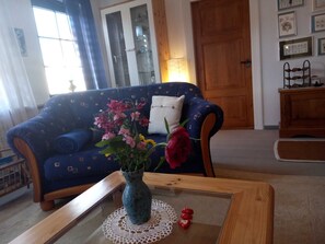 A living room with a blue patterned sofa, a glass coffee table with a lace doily, a vase with flowers, and a wooden cabinet.