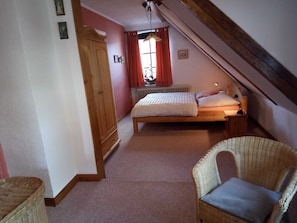 A bedroom with a bed, a wooden wardrobe, a wicker chair, and a window with red curtains. - Apartment Käding in Kappeln an der Schlei - Apartment Käding in Kappeln an der Schlei (Kappeln)