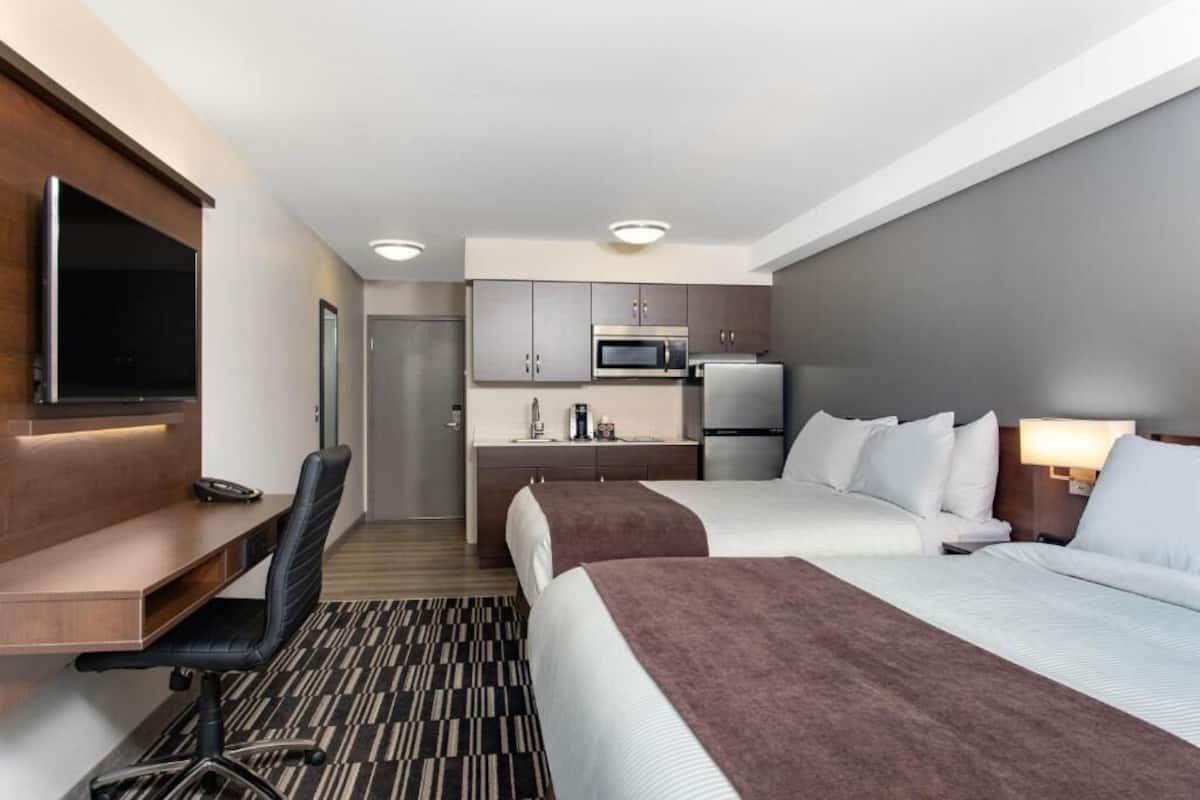 A hotel room with two beds, a desk, a TV, a kitchenette, and a bathroom.