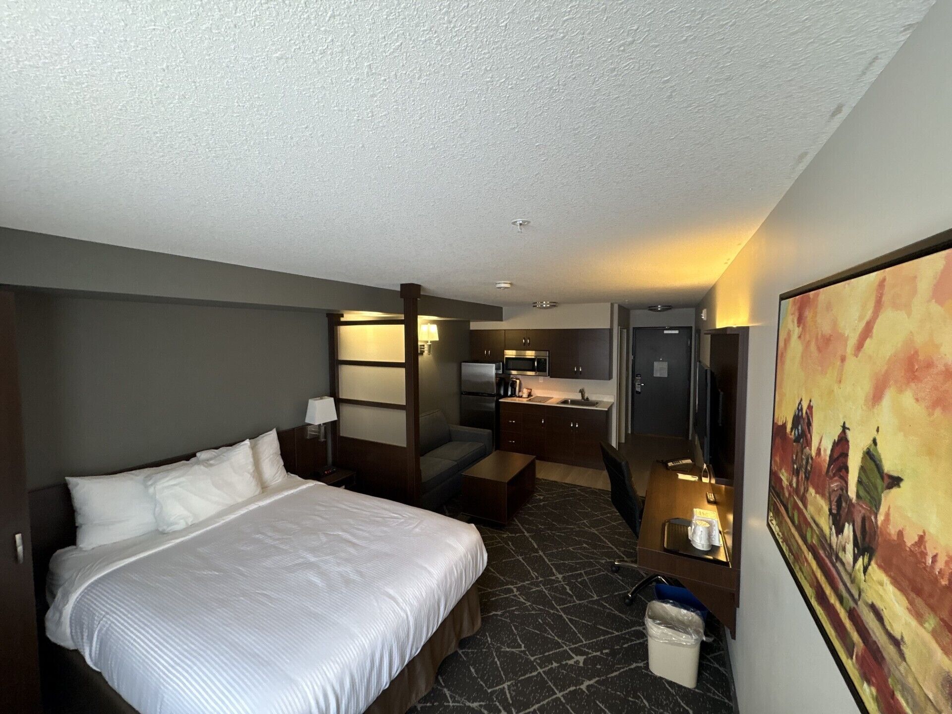 A hotel room with a large bed, a desk, a chair, a small kitchenette, and a painting on the wall.