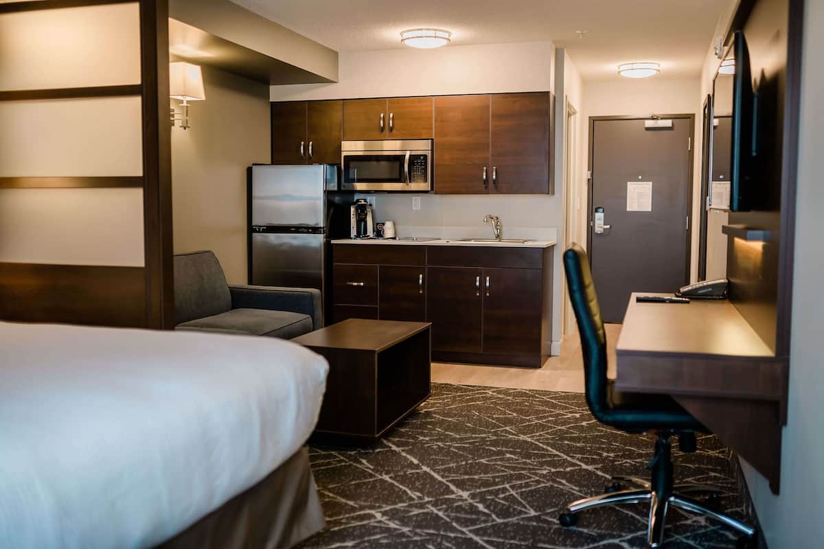 A compact hotel room with a bed, a small sofa, a desk with a chair, a kitchenette with a microwave and refrigerator, and a bathroom with a mirror and door.