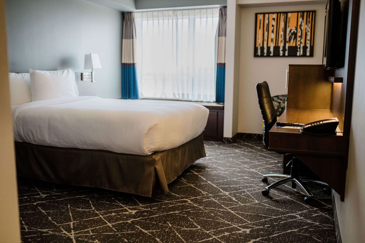 A hotel room with a large bed, a desk with a chair, a lamp, and a window with curtains.