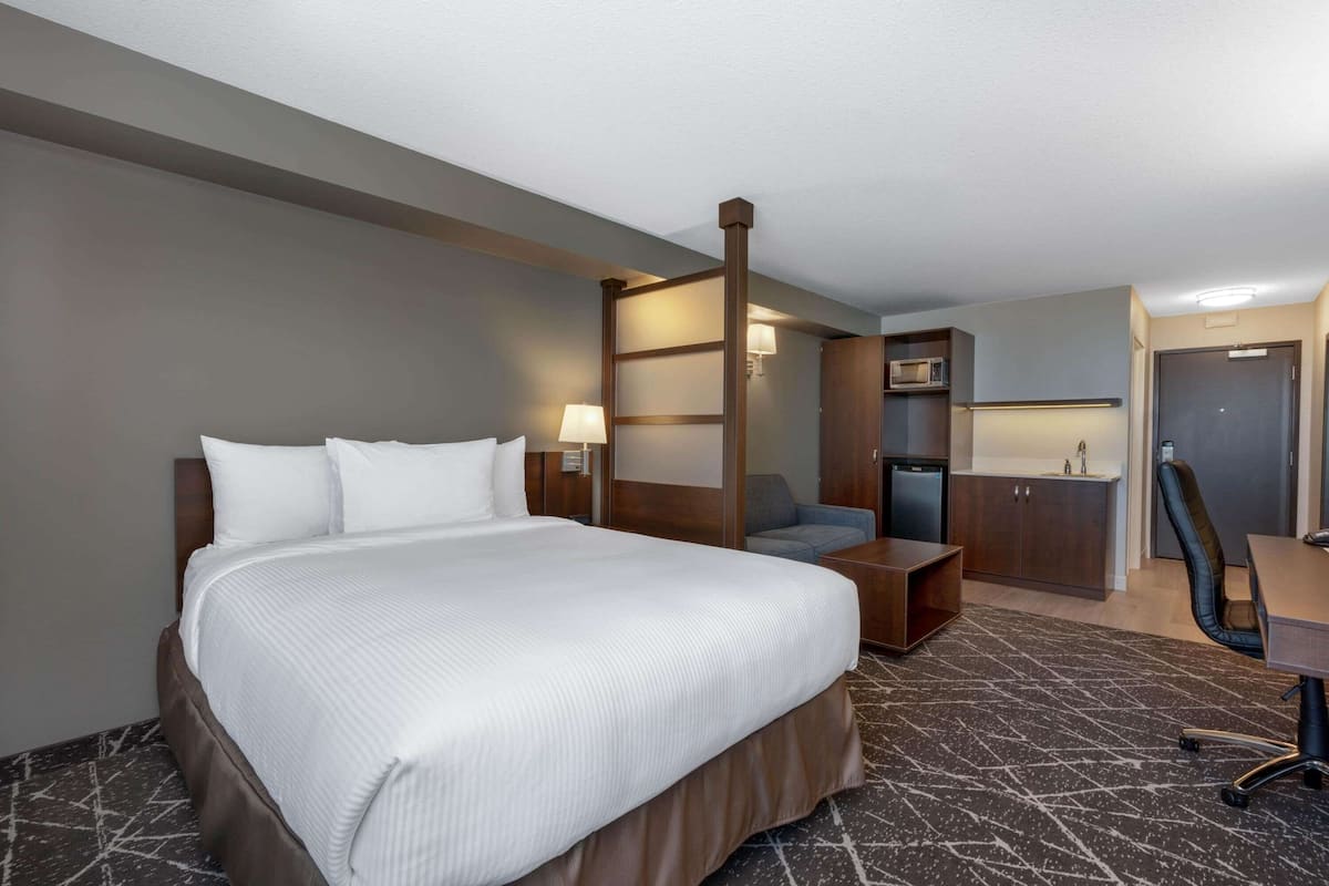A hotel room with a large bed, a desk with a chair, a microwave, a small kitchenette, and a door leading to another room.