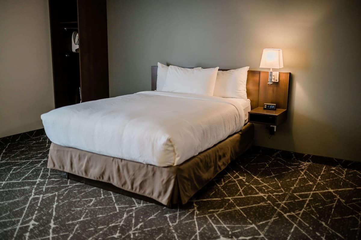 A neatly made bed with a headboard, a bedside table with a lamp, and a clock. The room has a dark marble floor and a plain wall.