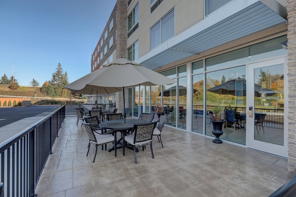 An outdoor patio with tables and chairs, a large umbrella, and a glass door leading to an interior space. - Holiday Inn Express & Suites Auburn Downtown by IHG (Auburn)