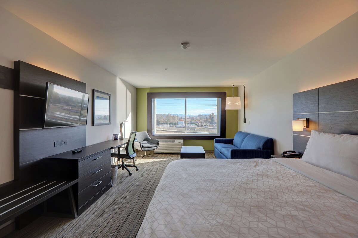 A hotel room with a large bed, a desk with a chair, a TV, a window with a view, and a lamp.